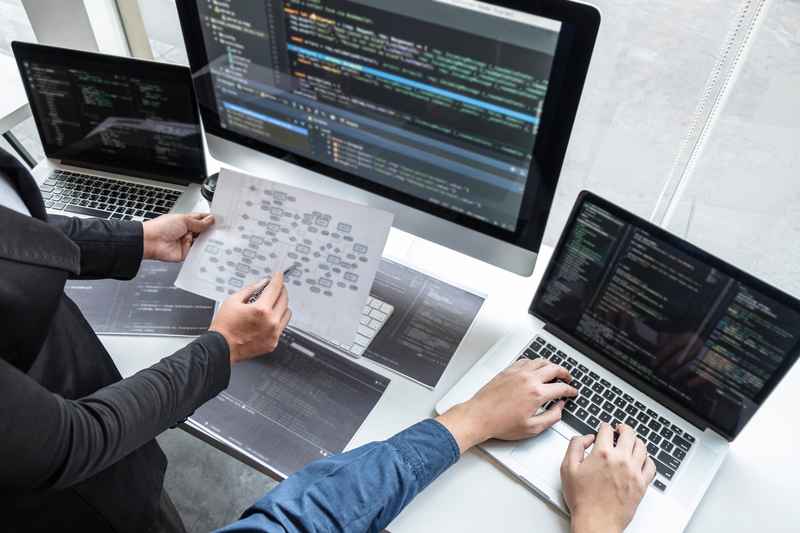 Vielfältiges Team junger Fachleute, die an einem modernen Büroschreibtisch mit Laptops und Tablets zusammenarbeiten. Sie sind in Diskussionen vertieft, zeigen auf Bildschirme und tauschen Ideen aus. Der helle, offene Arbeitsbereich schafft eine energiegeladene und kollaborative Atmosphäre, die Teamarbeit und digitale Innovation hervorhebt.